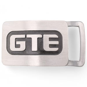 May include: A silver belt buckle with a black rectangular inset featuring the text "GTE" in a bold, sans-serif font.