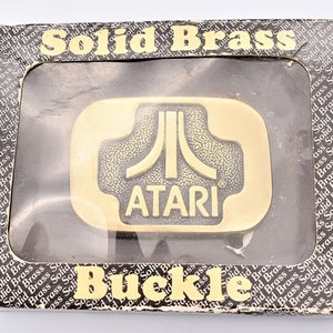 May include: A gold-colored metal belt buckle with the Atari logo and the word "ATARI" in black lettering. The buckle is in a clear plastic display case with a black background and the words "Solid Brass Buckle" printed on the box.