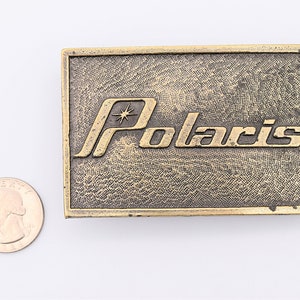 Puede incluir: Hebilla de cintur&oacute;n rectangular de lat&oacute;n con la palabra "Polaris" en letras en relieve. La hebilla tiene un fondo texturizado y un dise&ntilde;o de estrella encima de la "o" en Polaris. Una moneda de veinticinco centavos estadounidense est&aacute; al lado de la hebilla para comparar el tama&ntilde;o.