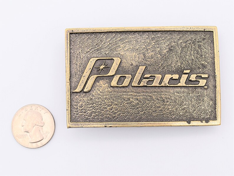 Puede incluir: Hebilla de cintur&oacute;n rectangular de lat&oacute;n con la palabra "Polaris" en letras en relieve. La hebilla tiene un fondo texturizado y un dise&ntilde;o de estrella encima de la "o" en Polaris. Una moneda de veinticinco centavos estadounidense est&aacute; al lado de la hebilla para comparar el tama&ntilde;o.