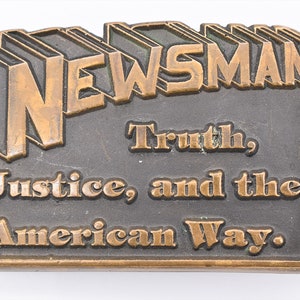 May include: A rectangular metal belt buckle with the word "NEWSMAN" in raised letters at the top. Below, the text reads "Truth, Justice, and the American Way." The buckle has a bronze finish.