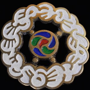 May include: A round, vintage brooch with a gold-tone metal frame and white enamel detailing. The center features a colorful design with red, green, and blue accents. The brooch has a detailed, ornate design, perfect for adding a touch of vintage flair.
