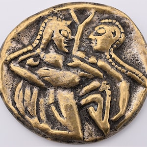 May include: A gold-colored metal coin with a raised design of two figures facing each other. The figures are both wearing elaborate headdresses and appear to be in a close embrace.