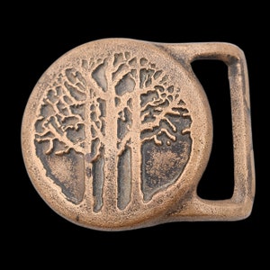 May include: A bronze-colored belt buckle with a circular design featuring a detailed tree silhouette. The buckle has a rectangular loop for the belt strap and a classic, rustic aesthetic. The tree design is intricately carved.
