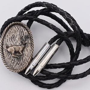 May include: A black leather bolo tie with a silver-toned oval pendant featuring a deer in a forest scene. The pendant is attached to two silver-toned bullet casings.