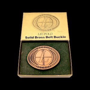 May include: A solid brass belt buckle in a box. The buckle is oval-shaped with the word "LEUPOLD" and "SIGHTS" around a circular design. The box is gold-colored with the words "LEUPOLD Solid Brass Belt Buckle". The buckle is bronze-colored and sits on a green felt lining.