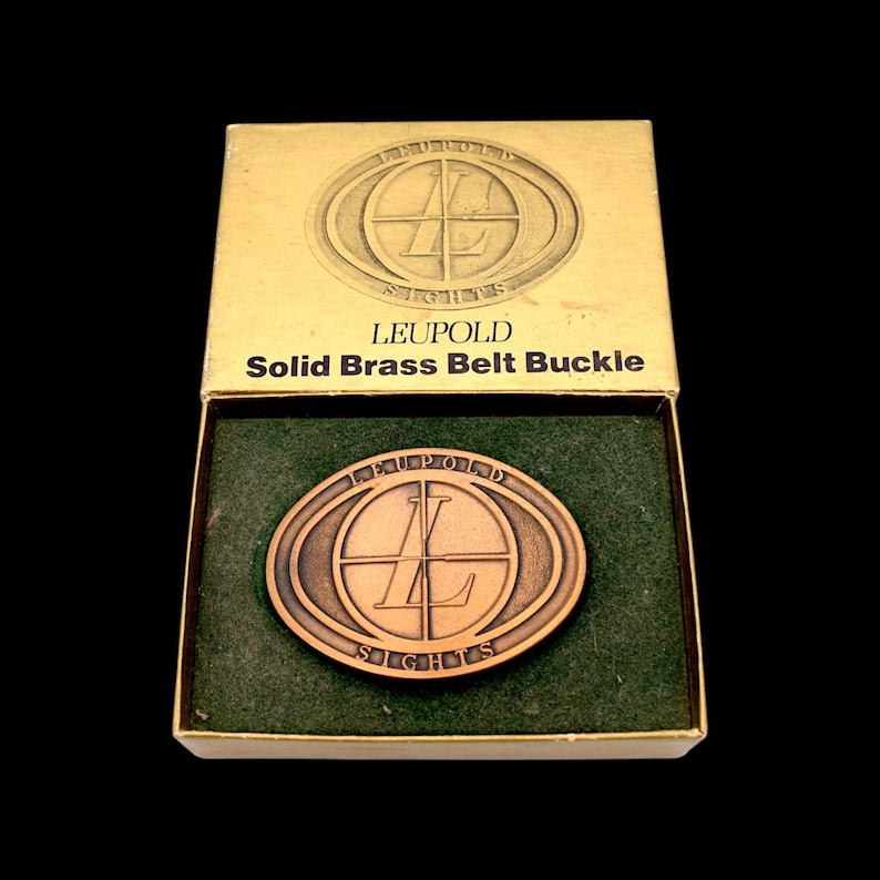 May include: A solid brass belt buckle in a box. The buckle is oval-shaped with the word "LEUPOLD" and "SIGHTS" around a circular design. The box is gold-colored with the words "LEUPOLD Solid Brass Belt Buckle". The buckle is bronze-colored and sits on a green felt lining.