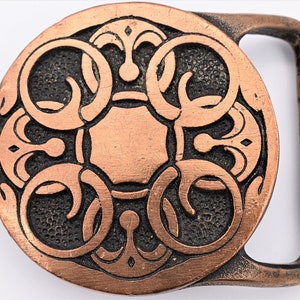 May include: A copper-colored belt buckle with an ornate, circular design. The buckle features a central square surrounded by swirling patterns and decorative elements. The metal has a slightly aged appearance.