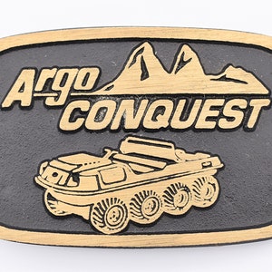 May include: A rectangular belt buckle with a black background and gold-colored trim. The buckle features the words "Argo CONQUEST" and an illustration of an amphibious vehicle. The mountains are also in gold.