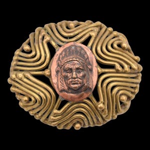 May include: A gold-toned metal brooch with a copper-toned oval medallion featuring a raised image of a Native American man's face. The medallion is surrounded by a decorative design of wavy lines.