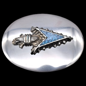 May include: Oval silver-toned belt buckle with an arrowhead design. The arrowhead features a turquoise inlay and detailed metalwork. The buckle has a polished finish and is set against a black background.