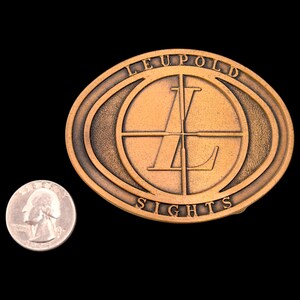 May include: Bronze-colored oval belt buckle with the text "LEUPOLD SIGHTS" and a crosshair design. A U.S. quarter is next to the buckle for size comparison. The buckle has a vintage, antique appearance.