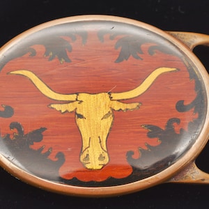 May include: A vintage oval-shaped belt buckle with a gold longhorn steer design on a red and black background.