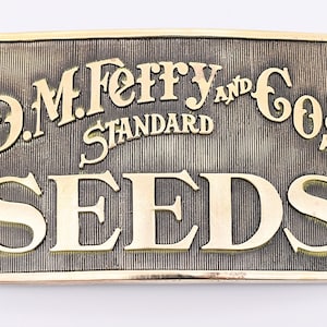 May include: A brass belt buckle with the text "D.M. Ferry & Co's Standard Seeds" in a gold-colored font.