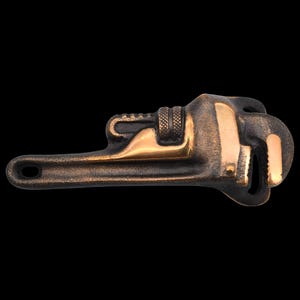 May include: A bronze-colored adjustable wrench with gold-colored accents. The wrench is a tool with a long handle, an adjustable jaw, and a hole at the end of the handle. The tool is set against a black background.