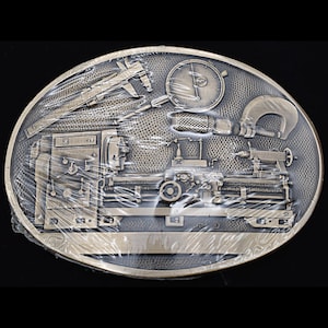 May include: A bronze belt buckle with a detailed image of a lathe, calipers, and a dial gauge. The buckle is oval shaped and has a textured surface.