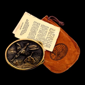 May include: An oval brass belt buckle with a detailed relief of two birds in flight over a landscape. The buckle is accompanied by a brown suede pouch and a tag with text describing the manufacturing process.