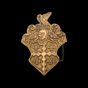 May include: A gold-colored metal belt buckle with a shield design. The shield features four crosses and a helmeted figure, topped with a bird. The buckle has a classic, vintage aesthetic.