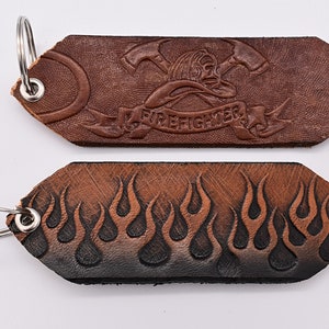 May include: Two leather keychains with metal rings. The top keychain is brown and features a firefighter helmet with crossed axes and the word "FIREFIGHTER". The bottom keychain is brown and black and features a row of flames.