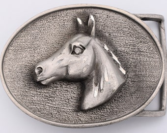 May include: A silver-toned metal belt buckle with an oval shape. The buckle features a detailed relief of a horse's head, with a textured background. The horse's mane and eye are clearly defined.