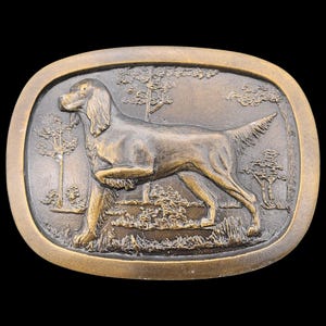 May include: A bronze-colored oval belt buckle featuring a detailed relief of a dog in a field with trees. The dog is in a hunting pose, with one paw raised. The buckle has a textured border and is set against a black background.