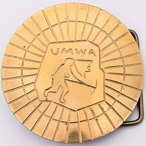 May include: A round, gold-colored belt buckle with a sunburst design. The center features a shield with the letters "UMWA" and a silhouette of a person. The buckle has a rectangular clasp.