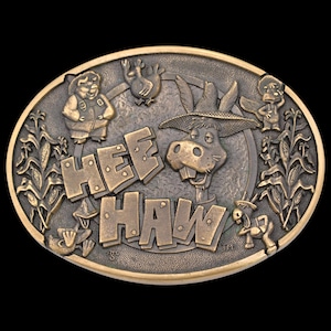 May include: Oval-shaped, bronze-colored belt buckle featuring the words "HEE HAW" in large, block letters. The buckle depicts cartoon characters, including a donkey wearing a straw hat, and other farm animals. Corn stalks frame the sides.