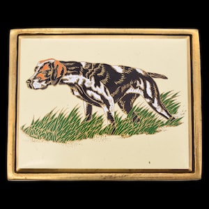 May include: Rectangular gold-toned belt buckle featuring a pointer dog illustration. The dog is depicted in black, white, and orange, standing in green grass against a cream background. The buckle has a gold border and is set against a black backdrop.