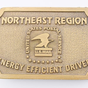 May include: A gold-colored metal belt buckle with the text "Northeast Region United States Postal Service Energy Efficient Driver" and a United States Postal Service logo.