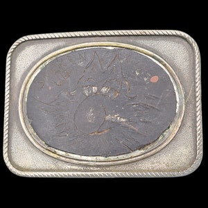 May include: A rectangular silver-toned belt buckle with a rope-like border. The center features an oval with a dark, textured surface, etched with a cartoon character and the words "SMOKIN' CLUB".