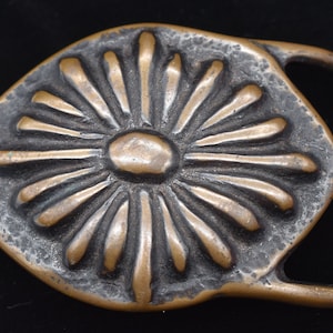 May include: A bronze-colored belt buckle with a sunburst design. The buckle has a rectangular loop and a central, raised sun-like pattern with radiating lines. The metal has a textured, aged appearance.