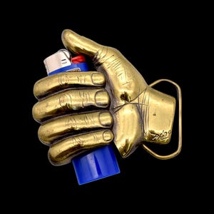 May include: A brass-colored belt buckle in the shape of a hand holding a blue and white lighter. The buckle has a rectangular frame and a detailed depiction of a hand with visible knuckles and lines. The lighter is held firmly in the hand.