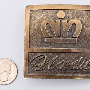 K&ouml;nnte beinhalten: Eine goldfarbene G&uuml;rtelschnalle aus Metall mit einer erhabenen Krone und dem darunter eingravierten Wort "Nordic".