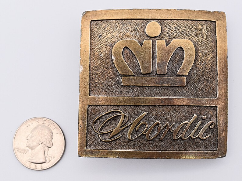 K&ouml;nnte beinhalten: Eine goldfarbene G&uuml;rtelschnalle aus Metall mit einer erhabenen Krone und dem darunter eingravierten Wort "Nordic".