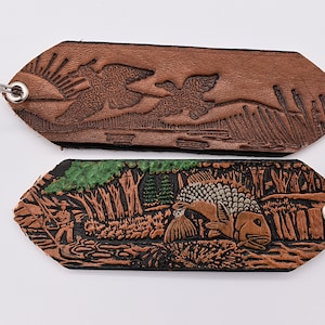 May include: Two leather keychains with embossed designs. The top keychain features a brown leather background with two birds flying over a river and reeds. The bottom keychain features a black leather background with a green tree and a fish in a river.