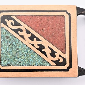 May include: A rectangular belt buckle with a handle. The buckle features a light brown wooden frame with a black border. Inside, a diagonal design divides the space into a red section and a turquoise section with a black decorative element.