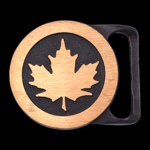 May include: A round, bronze-colored belt buckle with a black background. The buckle features a maple leaf design in the center, surrounded by a bronze ring. The buckle has a rectangular bar for the belt.
