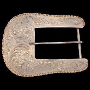 May include: Silver-colored belt buckle with floral and scroll engraving. The buckle has a rectangular opening and a gold-colored rope trim around the edges. The design features detailed floral patterns and a classic western aesthetic.