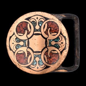 May include: A round copper-colored belt buckle with an ornate design. The buckle features a central square and circular patterns filled with red and turquoise-colored stones, outlined in black. The buckle has a dark, rectangular frame.