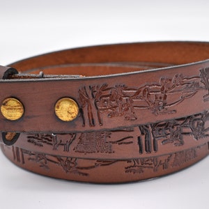 May include: A brown leather belt with a detailed embossed design of trees and animals. The belt has two gold-colored metal rivets.