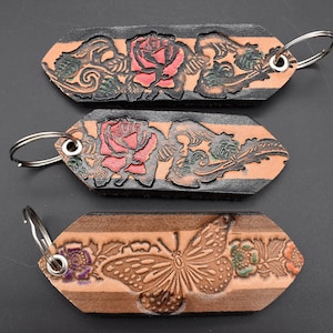 May include: Three leather keychains with different designs. The first keychain features a black leather background with a red rose and green leaves. The second keychain features a brown leather background with a red rose and green leaves. The third keychain features a brown leather background with a butterfly and green leaves.