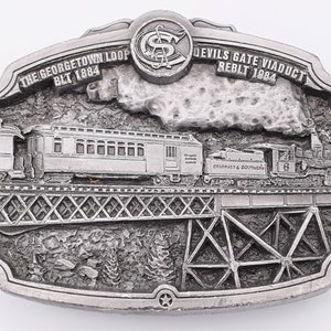May include: A silver-toned belt buckle featuring a train crossing a bridge. The train is labeled "Colorado & Southern" and the bridge is labeled "The Georgetown Loop Blt 1884" and "Devils Gate Viaduct Reblt 1984".