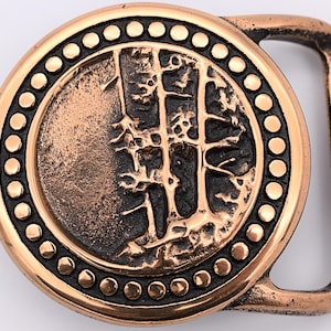 May include: A copper-toned belt buckle with a circular design. The center of the buckle features a raised, textured image of three trees. The outer edge of the buckle is decorated with a row of small, round dots.