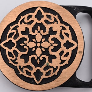 May include: A decorative belt buckle with a circular copper-colored center featuring an ornate, symmetrical design. The design is set against a black background, and the buckle has a black, rectangular frame.