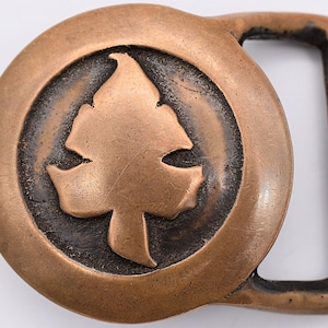 May include: A bronze belt buckle with a stylized leaf design in the center. The buckle is round and has a smooth, polished finish.
