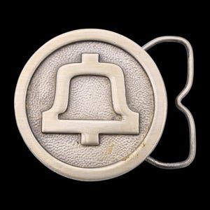 May include: A silver-colored belt buckle with a bell design inside a circular frame. The buckle has a textured background and a curved metal piece for attaching to a belt. The bell is a simple, stylized shape.