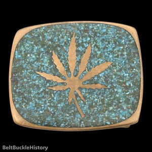 May include: A rectangular belt buckle with rounded corners featuring a central brass-toned cannabis leaf inlay. The background is composed of a mosaic of small, crushed turquoise and dark green stone fragments set within a metallic frame.