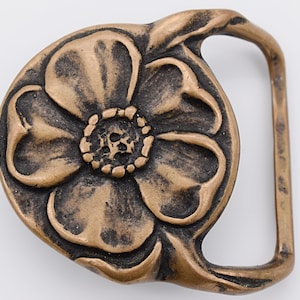 May include: A bronze belt buckle with a stylized flower design. The buckle is in the shape of a flower with five petals and a center.