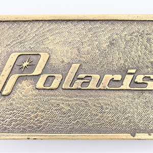Puede incluir: Hebilla de cintur&oacute;n rectangular de estilo vintage con la palabra "Polaris" en una fuente dorada estilizada. La hebilla tiene una superficie texturizada de color bronce y un gr&aacute;fico de estrella sobre la palabra "Polaris".
