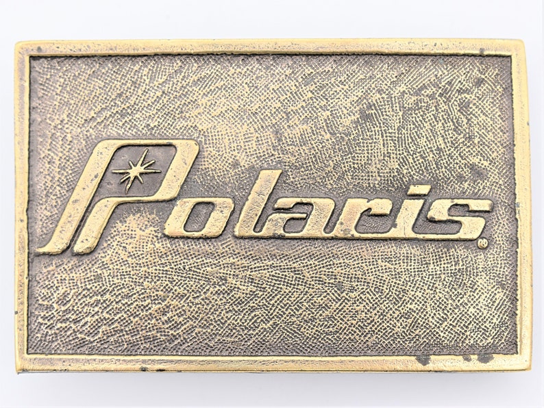 Puede incluir: Hebilla de cintur&oacute;n rectangular de estilo vintage con la palabra "Polaris" en una fuente dorada estilizada. La hebilla tiene una superficie texturizada de color bronce y un gr&aacute;fico de estrella sobre la palabra "Polaris".